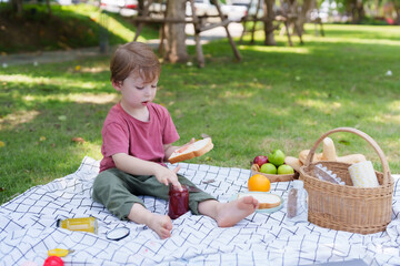 Adorable Caucasian boy is sitting on the grass at the village park, hungry, dipping his fingers...