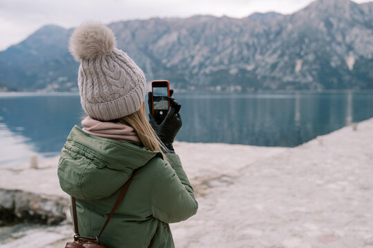 Woman stands on the seashore and takes pictures of the mountains on a smartphone. Back view