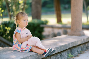 Little girl with a plush hare in her arms sits on a stone fence in the park and looks ahead