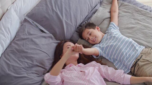 A smiling mom and son in playful and joyful mood lie in the bedroom on the bed. Demonstration the gesture of outstretched arms, reminiscent of airplane or scene from the movie Titanic. The concept of