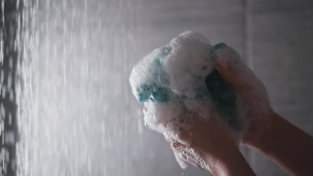 Woman squeezes loofah in shower cabin closeup. Lady hands hold foamy body washing sponge against water sprays. Soft supply for personal hygiene