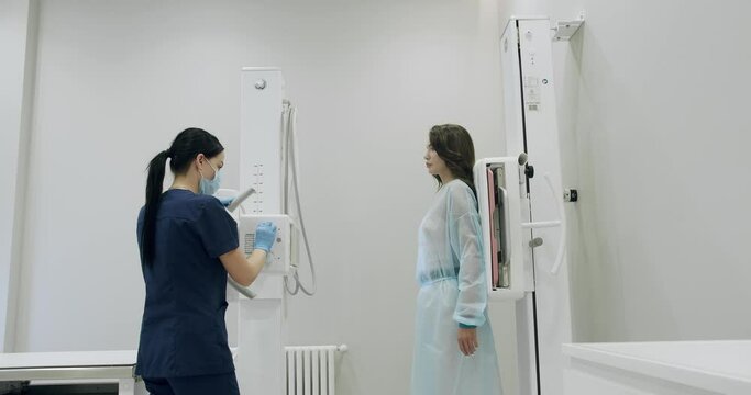 Woman patient in medical gown standing near x-ray machine in hospital. Nurse prepare for scanning female chest with modern medical equipment for examination in clinic. Health care concept
