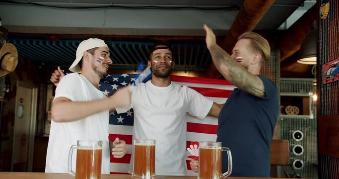 Man sports fan hold american flag in hands, watching match with friends together at pub. Joyful happy male with painted face giving high five to friend while cheering for football team in bar. 
