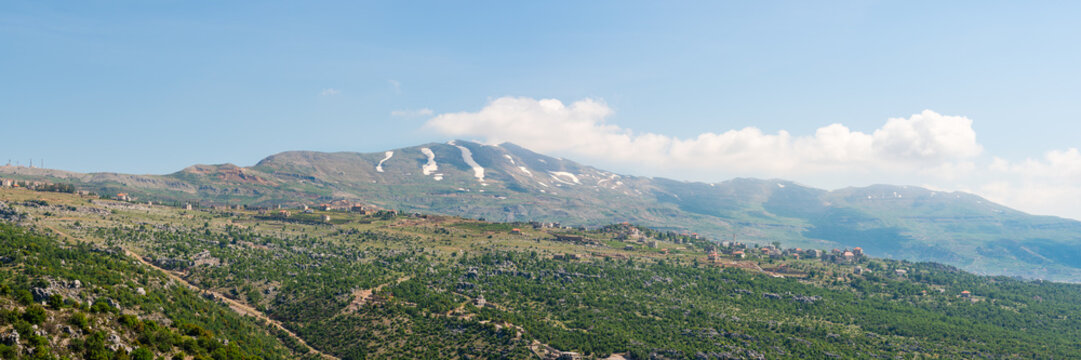 A village sits on a hill ridge across a rocky landscape, with Mount Lebanon mountain range, snow-covered in spring, a popular hiking destination in Lebanon, Middle East