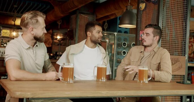 Multi ethnic serious men sitting in bar, drink beer and talking about life. Young males have conversation after work day at pub. Communication concept.