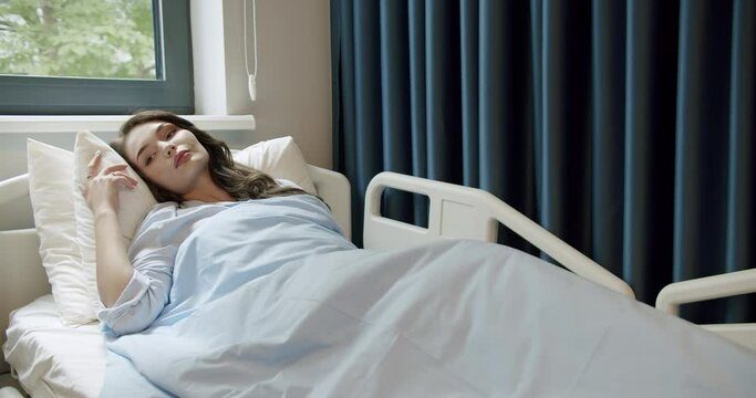 Young woman patient lying on comfortable bed in recovery room at hospital. Sick female resting in clinic ward room with modern interior. Medicare concept