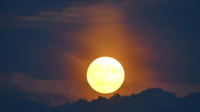 Dark blue sky and clouds illuminated in orange and pink around the rising yellow full moon - timelapse. Topics: weather, atmosphere, space, astronomy, dusk