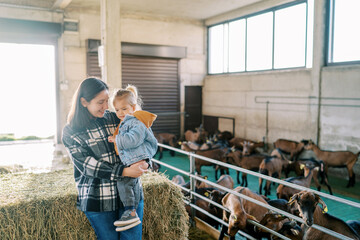 Mom with a little girl in her arms stands by a haystack near a goat paddock - obrazy, fototapety, plakaty