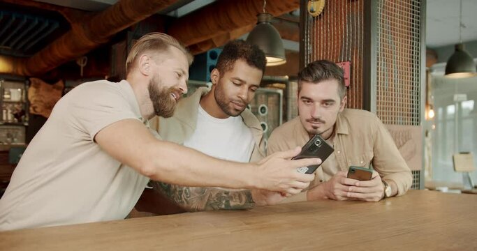 Men friends sitting and relax at bar with smartphones, talking and smile. Joyful male showing picture on cellphone to buddies in bar. Friendship and communication concept