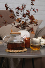 Autumn home design - hot teapot with tea, glass of tea, burning candle, vase with dry branches on sofa background in cozy living room.