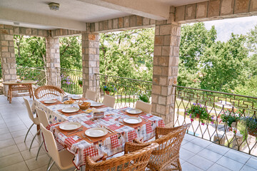 Laid festive table surrounded by rattan chairs stands on the terrace of a stone villa in a green...