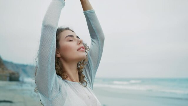 Woman in sheer blouse standing on ocean beach with raised hands, deeply breathing fresh ocean breeze on cloudy summer day. Freedom, relaxation, recovery, healing, meditation, nature concept 4k footage