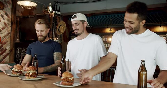 Multi ethnic men friends resting at weekend at pub, drinking beer, eating burgers and talking. Male buddies in casual clothes spending fun time together at bar after work and enjoy hamburgers