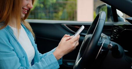 Smiling teen woman typing message on smartphone while sitting in car on drivers seat. Side view of...