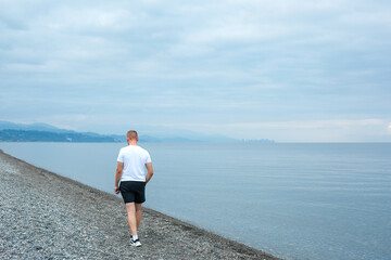 Fototapeta na wymiar A man in sportswear walks along a pebble beach by the sea