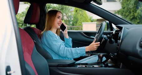 Naklejka premium Attractive ginger woman talks on smartphone while sits sits in car. Girl driver calling smartphone in modern automobile standing at sunny shore. Attractive blonde speaking on telephone inside auto.