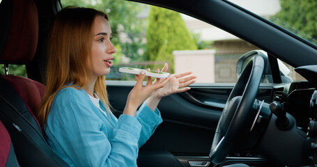 Focused blonde lady sitting behind the wheel of car when in recording voice message on smartphone...