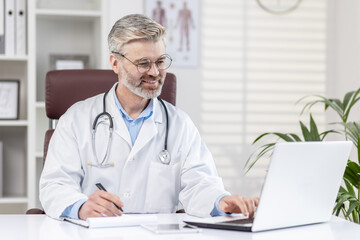 Mature experienced gray-haired doctor works remotely on paper work inside medical office inside clinic, doctor uses laptop at work, records data on patient.