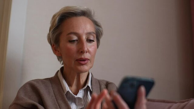 Closeup face of elegant middle-aged woman typing on mobile phone, using online application, media service, checking social media, wireless Internet technology at home, sitting on cozy sofa by window.