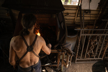 Back view of blacksmith stoking the fire in the forge of his workshop.