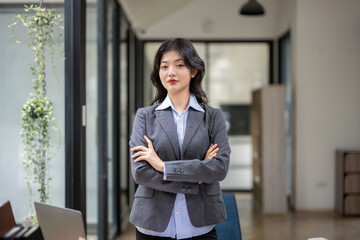 Obraz na płótnie Canvas Smart and self-confident Asian businesswoman standing with arms crossed at desk in office