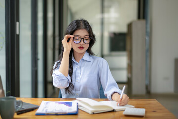 Asian businesswoman wearing glasses sits at her desk. Focus on writing in notebooks.