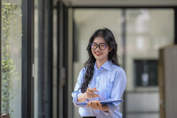 A disheveled businesswoman stands with happy smile wearing glasses and holding a clipboard. Thinking and writing a busy new project.