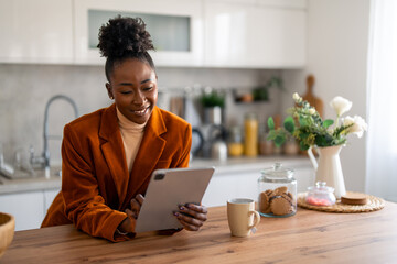 Smiling black woman entrepreneur working from home over digital tablet while standing at kitchen...