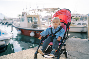 Little smiling girl sits in a stroller on the pier near the moored yachts and points to the sea