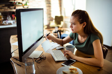 Young woman holding document looking at monitor with blank screen at home