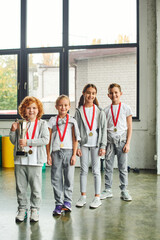 vertical shot of little boys and girls posing with golden medals and trophy, looking at camera