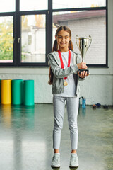 vertical shot of little cute girl with long hair with golden medal and trophy looking at camera