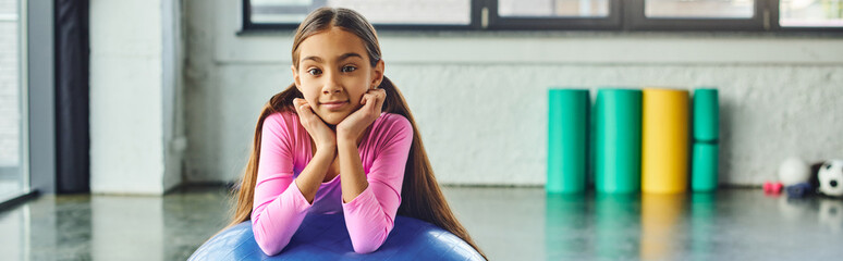 jolly little girl with long hair posing with fitness ball with hands under chin, child sport, banner