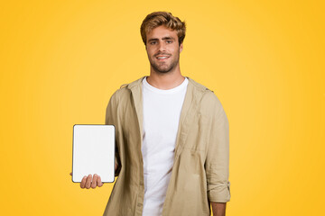 Joyful European man holding tablet with blank screen on yellow background