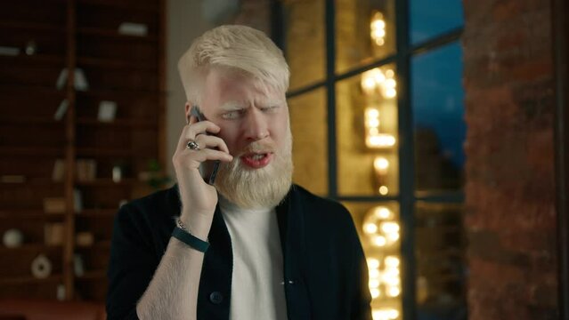 Smiling albino entrepreneur talking on smartphone in the loft office at night city. Portrait of casual businessman having phone conversation with coworker. Hipster man with surprised face expression 
