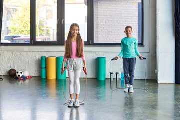 little cute boy and girl in sportswear posing with jump ropes and smiling at camera, child sport - obrazy, fototapety, plakaty