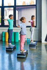three adolescent girls and boy in sportswear exercising on fitness stepper with arms asides, sport