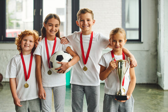 joyous little boys and girls with medals smiling at camera and holding trophy and football, sport