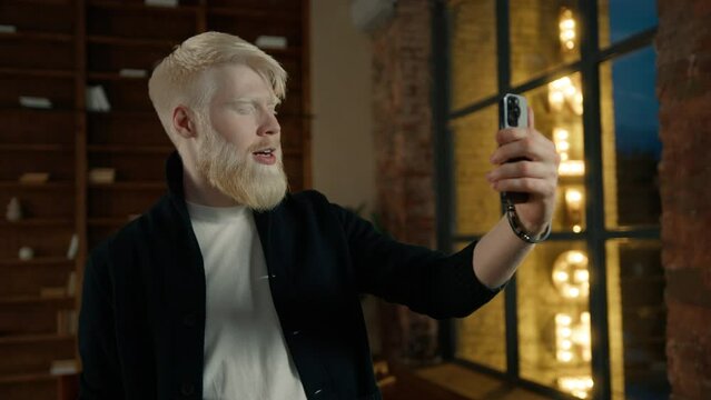 Casual albino businessman having video conversation on smartphone with street sign illuminated at night in window background. Male entrepreneur waving hand at camera. Smiling man in loft office night 