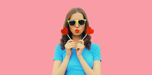 Portrait of beautiful young woman with red heart shaped lollipop blowing her lips with lipstick sending sweet air kiss wearing sunglasses on pink background - obrazy, fototapety, plakaty