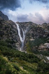 An incredible view of a majestic Shdugra waterfall, high in the Caucasus Mountains, rocky slopes, a rushing stream. Green herbs and plants with flowers. Mazeri, Svaneti, Georgia. Vertical photo