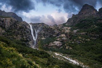 An incredible view of a majestic Shdugra waterfall, high in the Caucasus Mountains, rocky slopes, a rushing stream. Green herbs and plants with flowers. Mazeri, Svaneti, Georgia