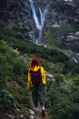 A woman, hiker walks along a path among high mountains. Shdugra waterfall in the background. Concept of hiking, active lifestyle, travel. Summer day, Georgia. Vertical photo