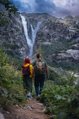 Back view of couple tourists, woman and man with backpack. Shdugra waterfall in the background. Concept of hiking, active lifestyle, travel. Vertical photo. Summer day, Mazeri, Georgia
