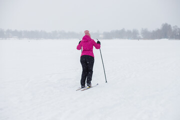 Skier in windbreaker and hat with ski poles in his hands with his back against the background of a snowy forest. Cross-country skiing on winter lake, outdoor sports, healthy lifestyle. Active people