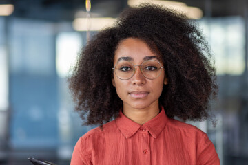 Close up portrait of young beautiful successful business woman, hispanic woman serious confident looking at camera close up, female worker inside office at workplace in red shirt with curly hair. - obrazy, fototapety, plakaty