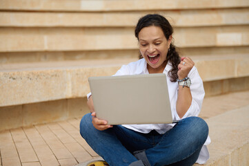 Young woman student with laptop, clenching fist, raising arm up, expressing positive emotion while passing exams