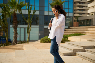 Happy young brunette female manager in casual attire, holding laptop and talking on mobile phone while walking outdoors