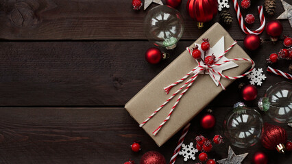 Gift wrapped in craft brown paper lies among holiday decor, red balls, stars, snowflakes and berries on wooden background, copy space, flat lay, top view.Merry Christmas,Happy New Year concept banner.