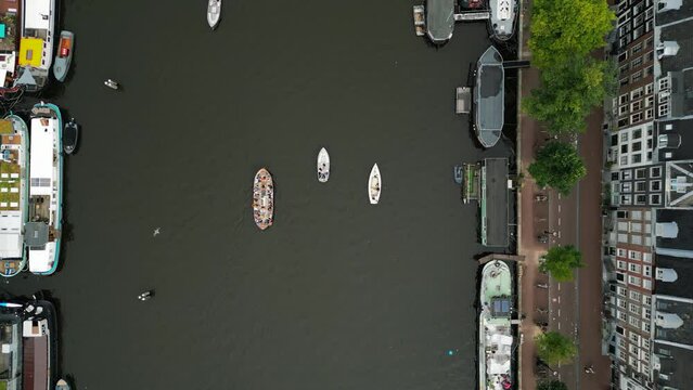 Drone footage of stunning view of boats and barges on narrow canal of Amsterdam with dense row of houses and roads with trees for pedestrians and cyclists. High quality 4k footage
